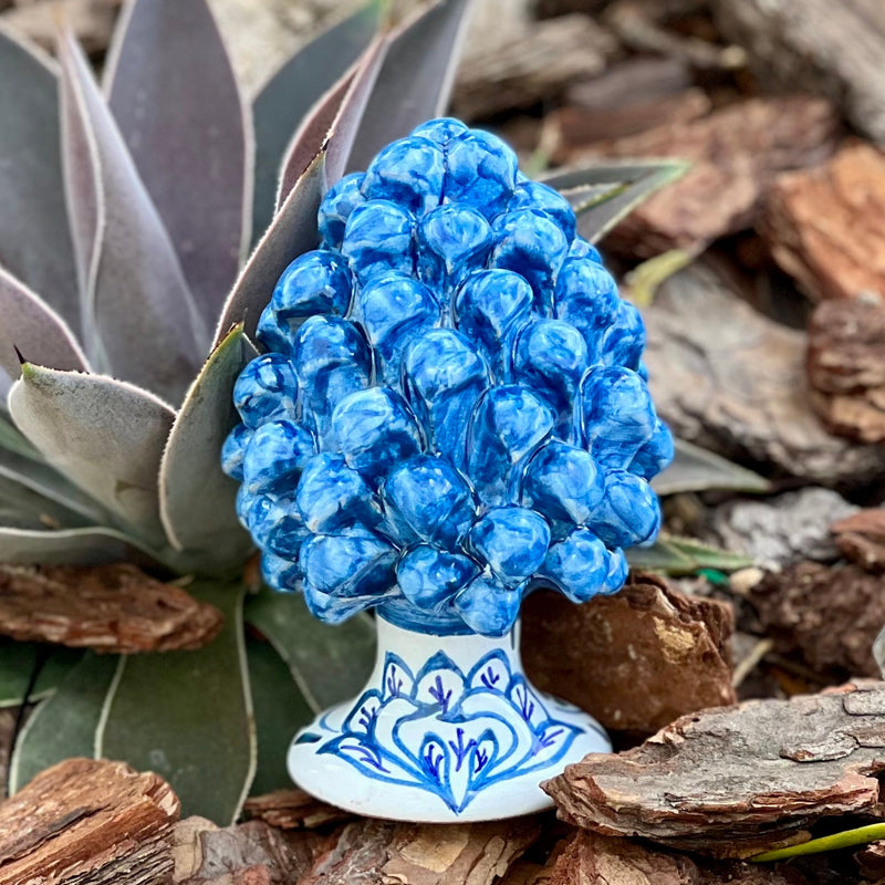 Blue ceramic hand painted pine cone on a natural background with green plant and wood pieces