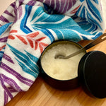 Black container with a spoo inserted in body scurb on a wooden surface with a Drifting Leaves towel in the background.