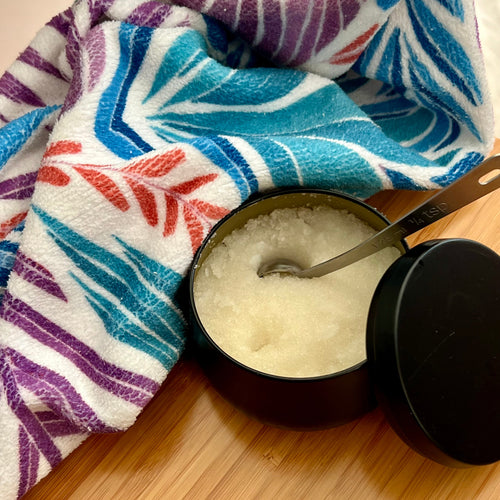 Black container with a spoo inserted in body scurb on a wooden surface with a Drifting Leaves towel in the background.
